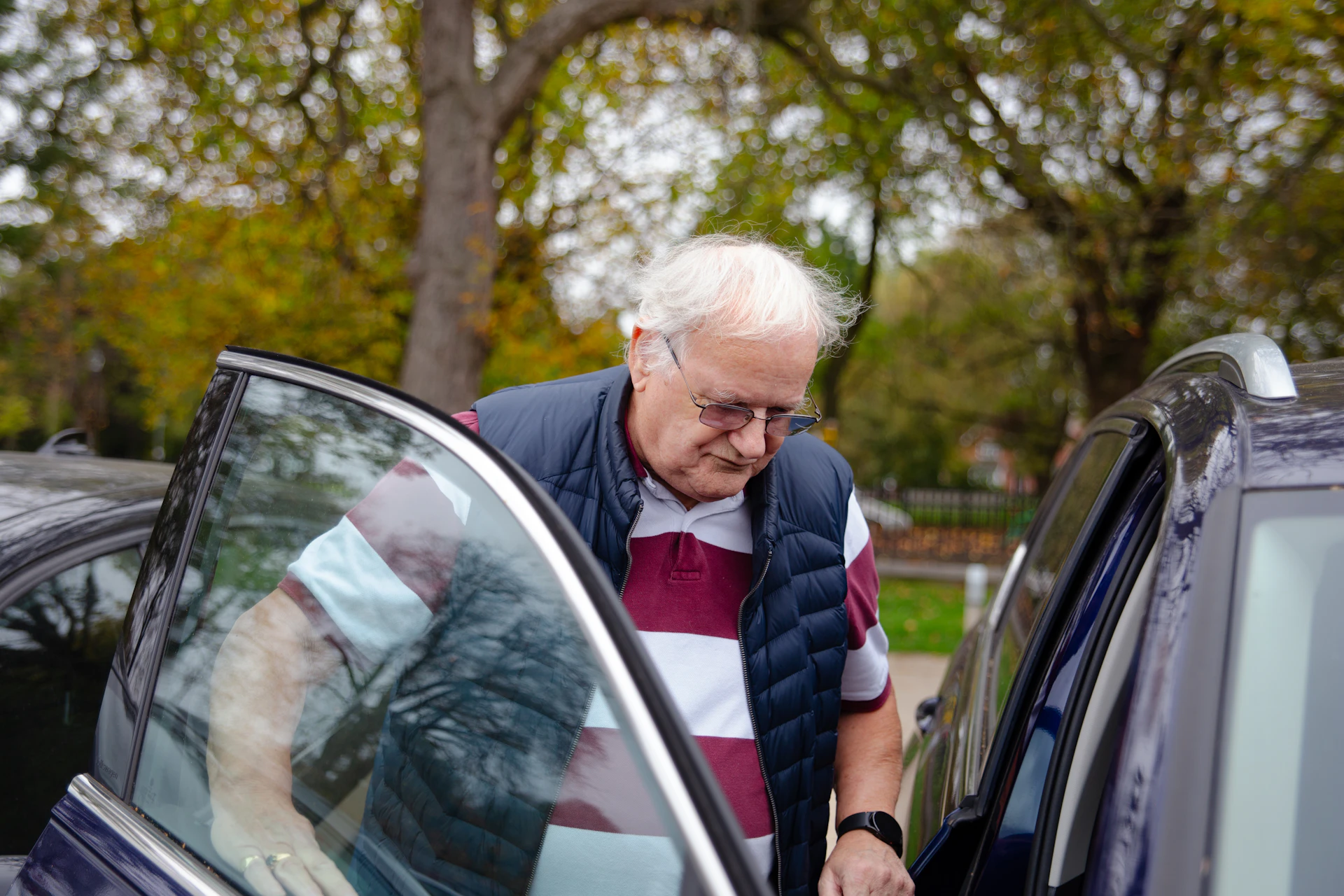 An elderly man being helped into a car by a caring driver in a quiet, tree-lined neighborhood